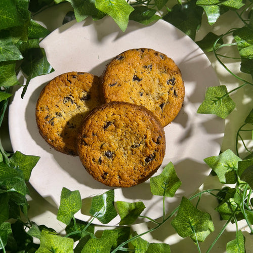 Three handcrafted chocolate chip cookies elegantly arranged on a white plate, styled with fresh green leaves.