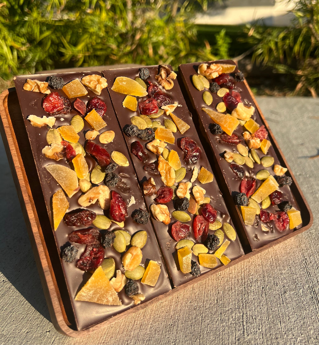 Three handcrafted chocolate bars featuring dried fruits and nuts, styled on a rustic wooden board in an outdoor setting.
