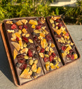 Three handcrafted chocolate bars featuring dried fruits and nuts, styled on a rustic wooden board in an outdoor setting.